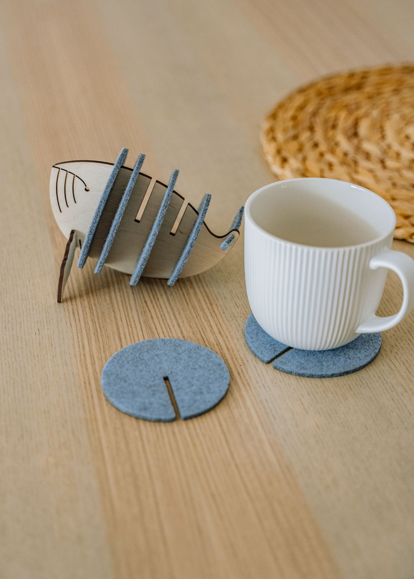 A white ribbed ceramic mug sits on a round gray felt coaster on a wooden table, with Whale Coasters and a whale-shaped holder nearby. A woven placemat adds home charm while these coasters help protect surfaces in style.