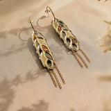 A pair of Wheat Statement Earrings with intricate cutout designs and dangling chain accents, displayed on a soft beige surface with delicate shadows and dried flowers—a bold design perfect for standout style.
