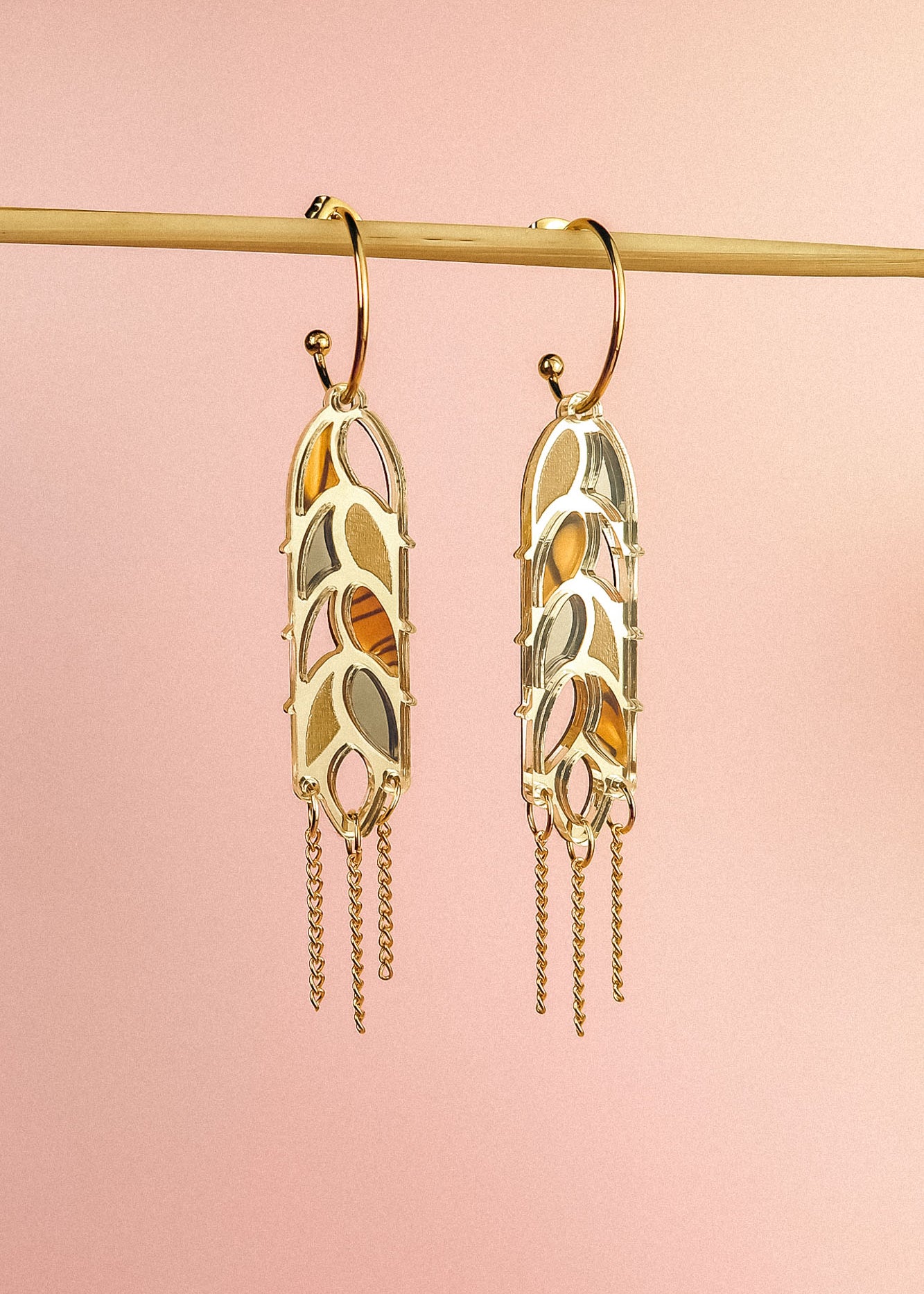 A pair of Wheat Statement Earrings with cut-out leaf patterns and dangling chain accents hang from a horizontal rod against a soft pink background, showcasing standout style.