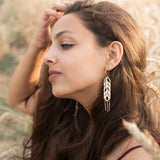 A young woman with long brown hair sits in a wheat field, wearing a burgundy dress and Wheat Statement Earrings. She gazes thoughtfully to the side, lightly touching her hair, her standout style enhanced by the bold design of her earrings.